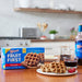 A kitchen table with Shop Wyman's Fruit First Waffles - Homestyle, a plate of waffles drizzled with syrup, strawberries, a glass of juice, and various kitchen items in the background. These individually wrapped waffles offer 10g of protein per waffle for a delicious and nutritious breakfast.