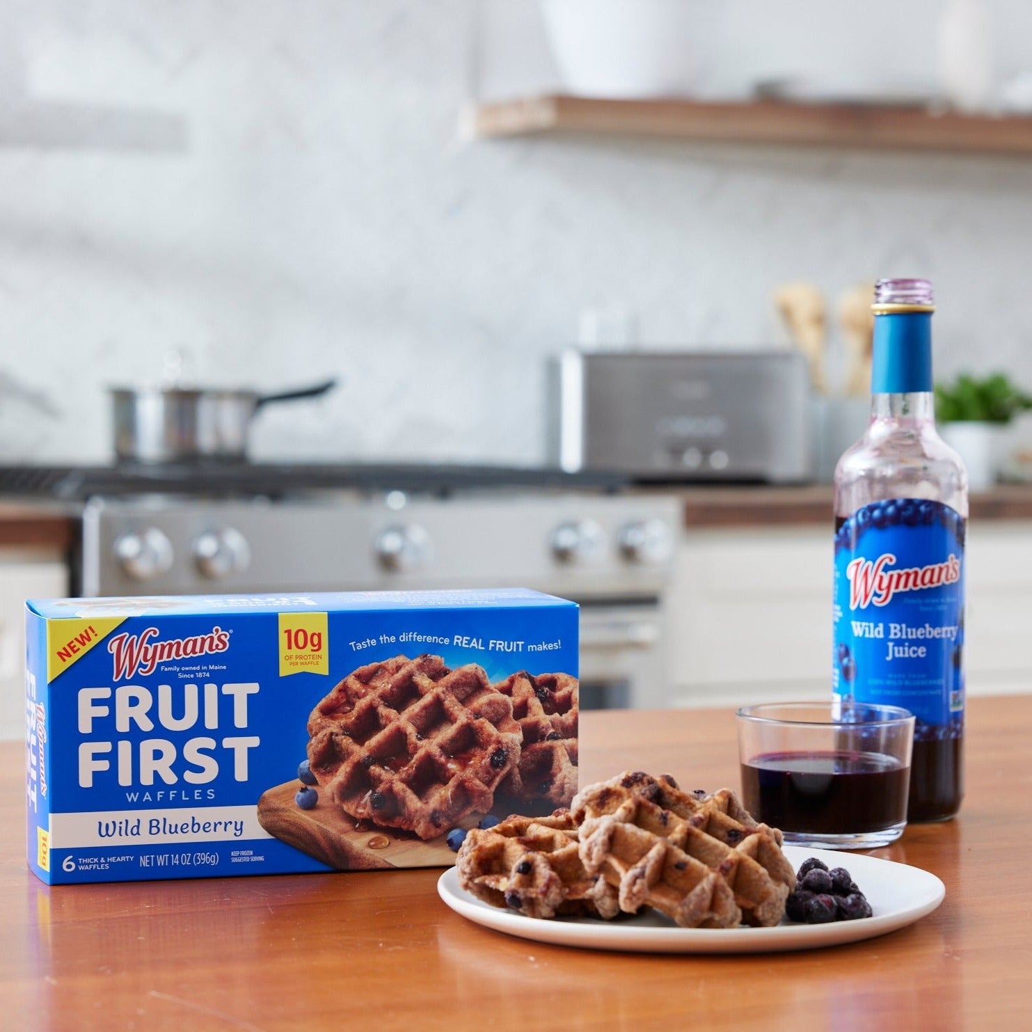 A box of Shop Wyman's Fruit First Waffles - Wild Blueberry and a bottle of Shop Wyman's Wild Blueberry Juice sit on a kitchen counter. A plate showcasing these high-protein breakfast waffles topped with wild blueberries and a glass of juice are ready to enjoy in front.