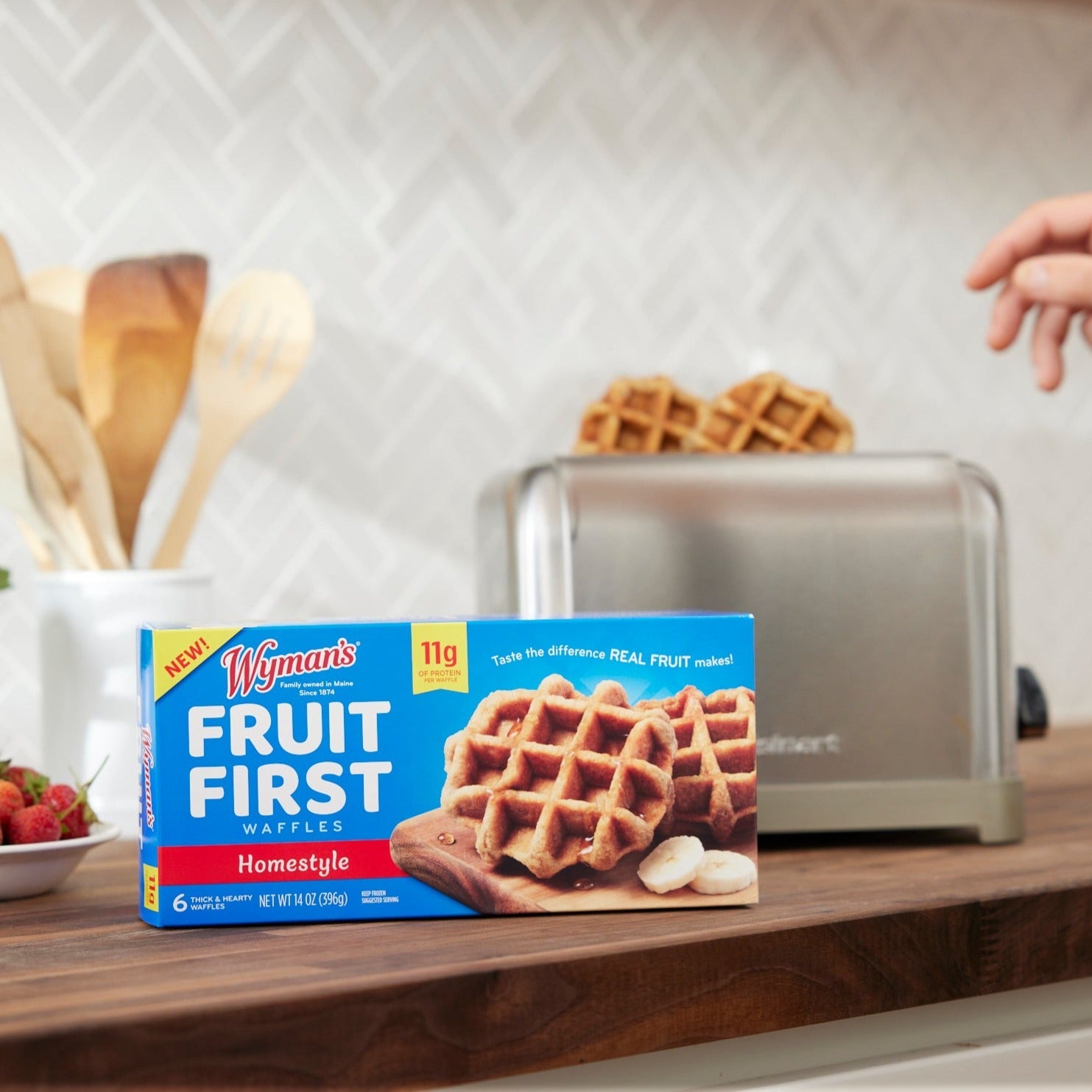 A box of Shop Wyman's Fruit First Waffles - Homestyle, boasting 10g of protein per waffle, is placed on a kitchen counter near a toaster with waffles being toasted and wooden utensils in the background.