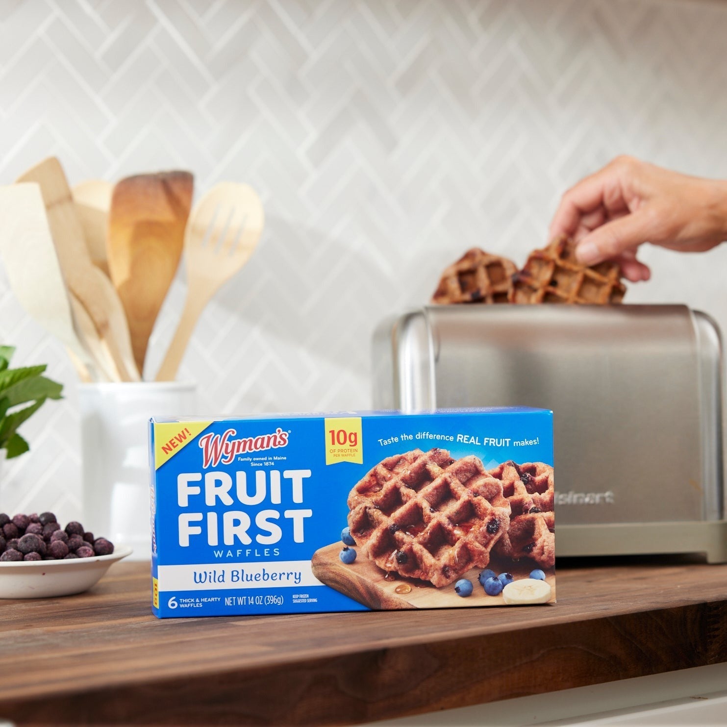 A box of Shop Wyman's Fruit First Waffles - Wild Blueberry sits on a kitchen counter as a hand places a waffle into the toaster. A bowl of wild blueberries and wooden utensils are visible in the background, all set for a high-protein breakfast.