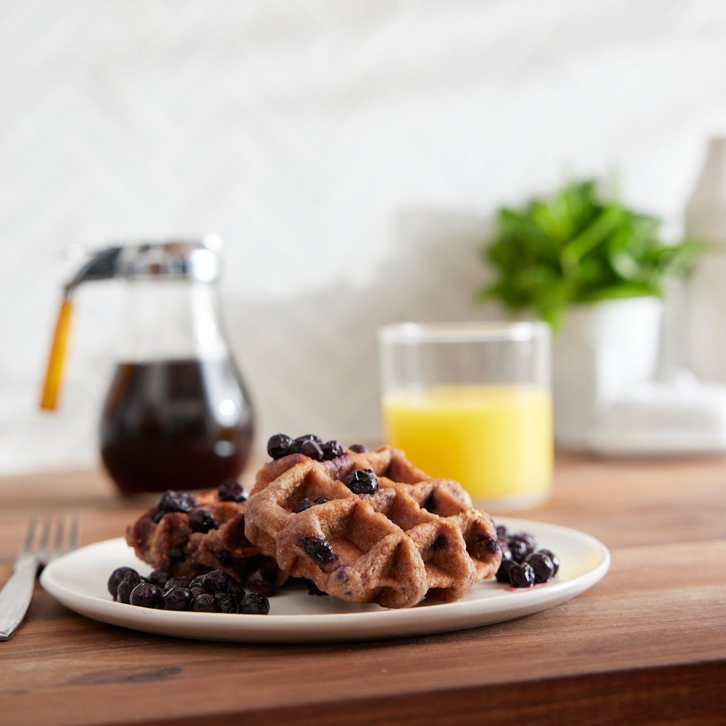 A plate of Shop Wyman's Fruit First Waffles - Wild Blueberry topped with wild blueberries sits on a wooden table with a glass of orange juice and a syrup dispenser in the background, making for the perfect high-protein breakfast.